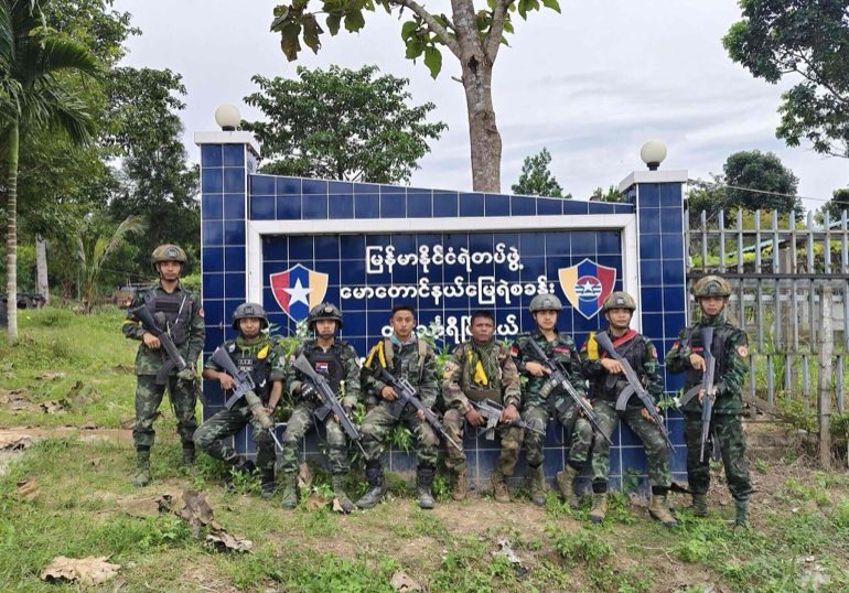 Armed men in camouflage uniforms and helmets posing with rifles in front of a Myanmar police station sign