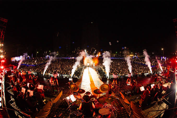 A night-time concert in Myanmar seen from behind the stage, with musicians in the foreground and a large crowd beyond